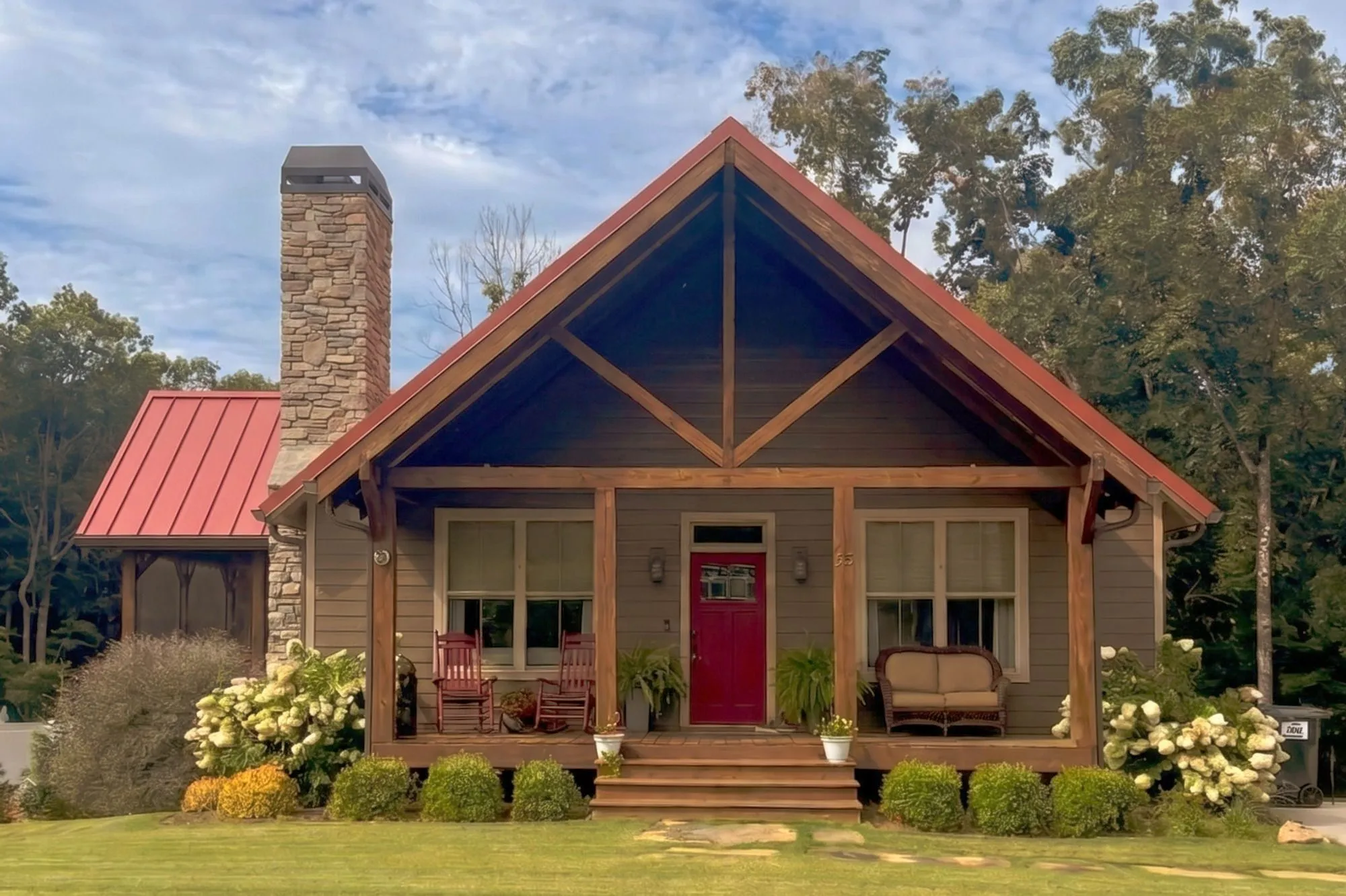 Exterior front view of The Soque — timber-frame porch gable, red standing-seam roof, stone-wrapped chimney, and hydrangeas along the foundation